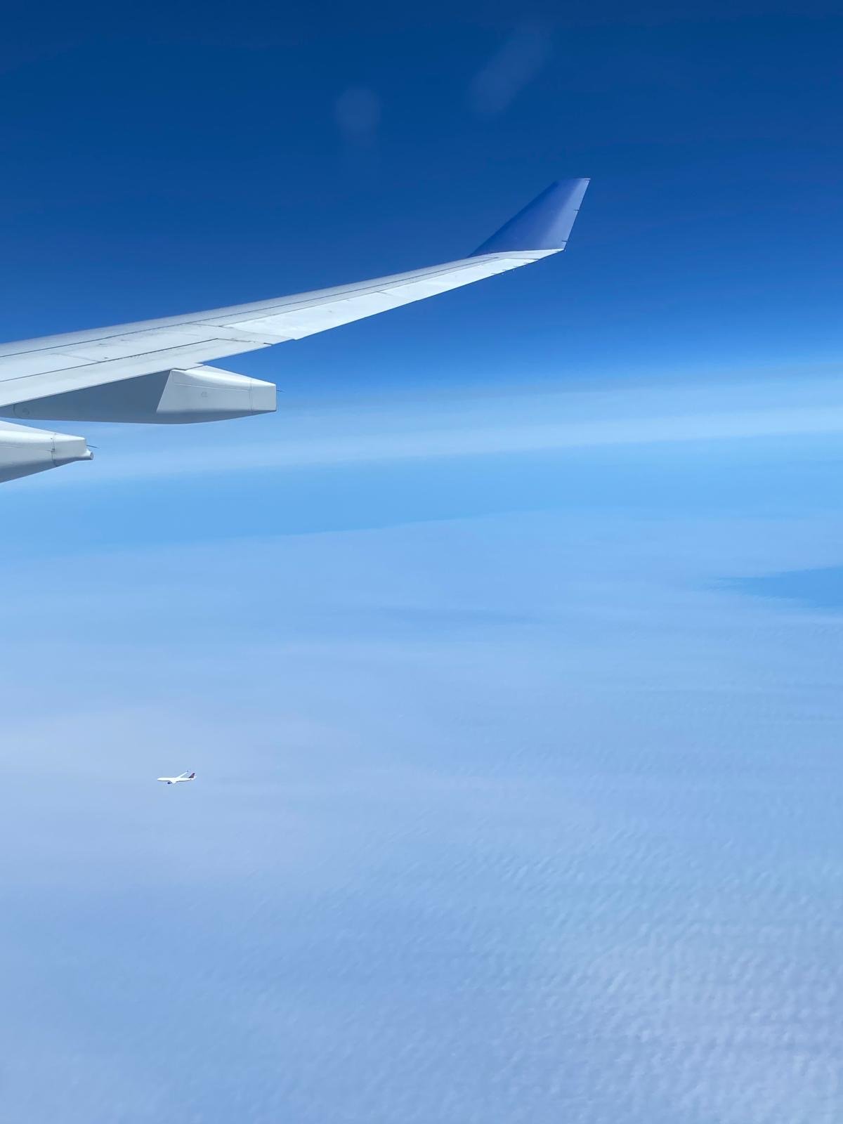 View from an airplane window showing the wing above the clouds and a second aircraft in the distance.