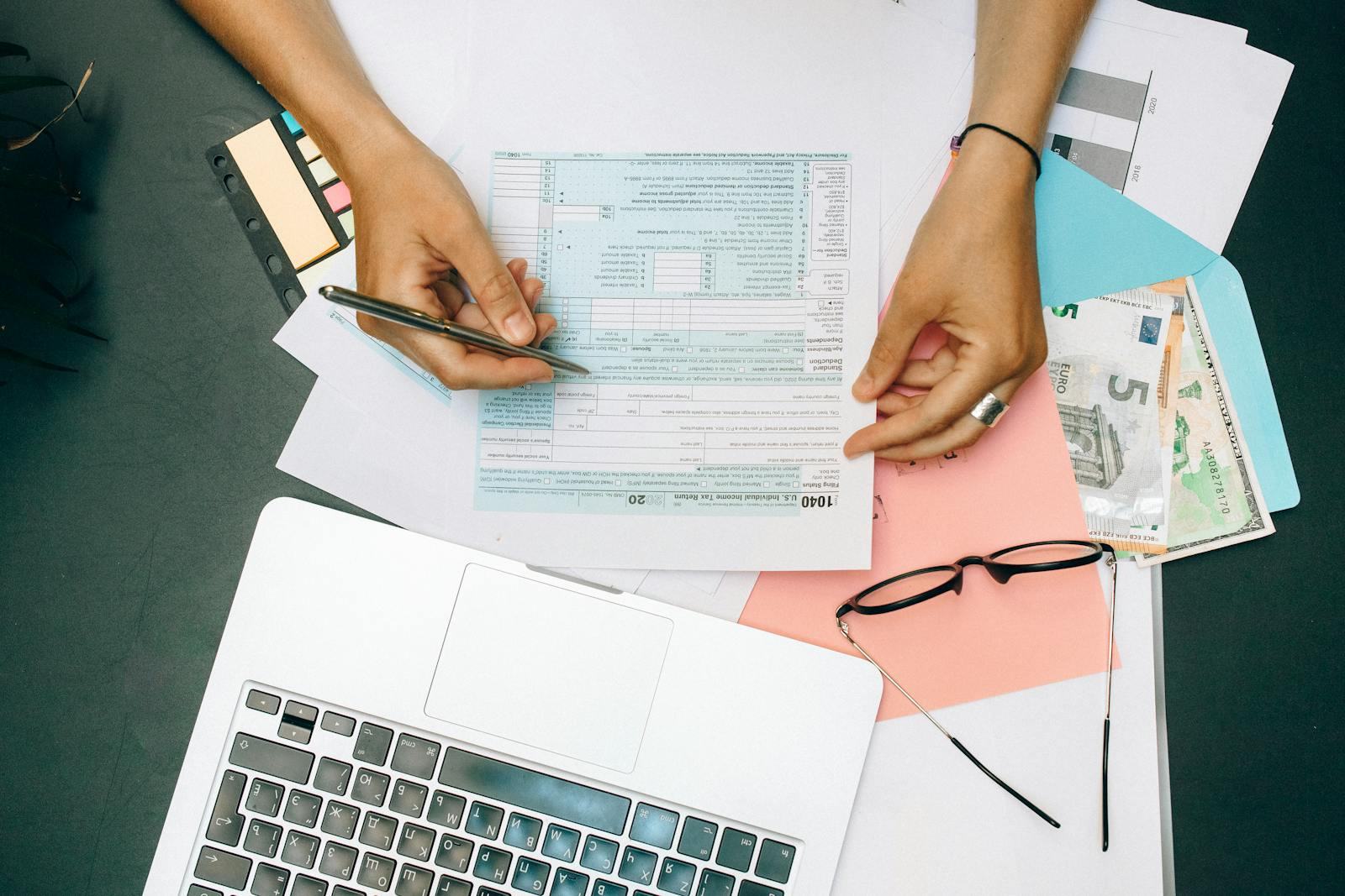Hands writing on tax documents at a desk with a laptop, eyeglasses, and currency, illustrating income-tax preparation work.