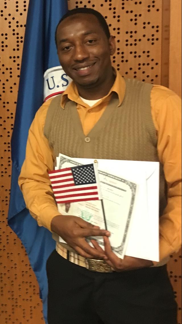 A new US citizen holding their naturalization certificate and a small American flag, standing in front of a US flag.